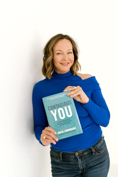 Anese Cavanaugh in a blue top holding up her book Contagious You, smiling at the camera
