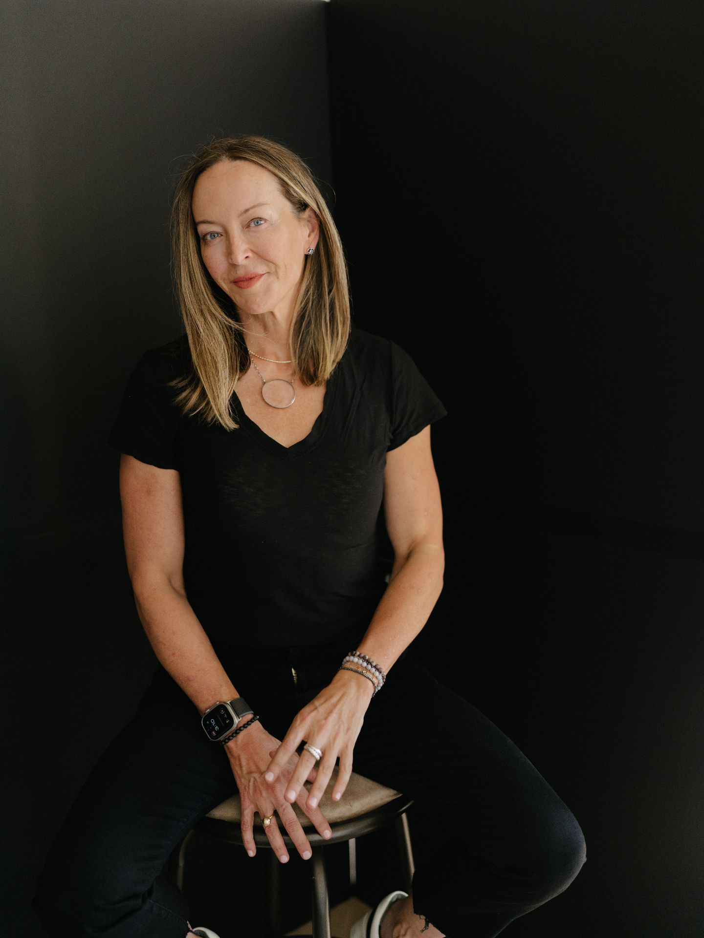 Anese Cavanaugh seated on a stool in a black v-neck against a dark backdrop, hands resting, direct gaze