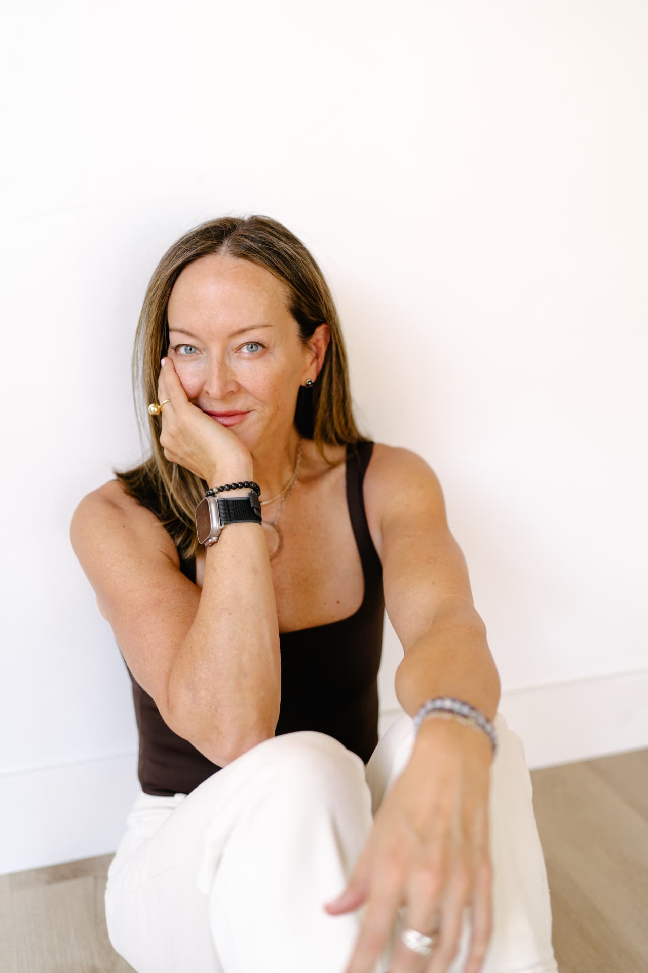 Anese Cavanaugh seated against a white wall in a brown tank top, chin resting on her hand, smiling warmly